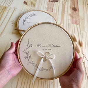 May include: Two round, beige ring holders with wooden frames. One features the names "Marine & Stephane" and the date "12.09.2026". The other has the name "Marjorie" and a date. Both have floral designs and a ribbon.