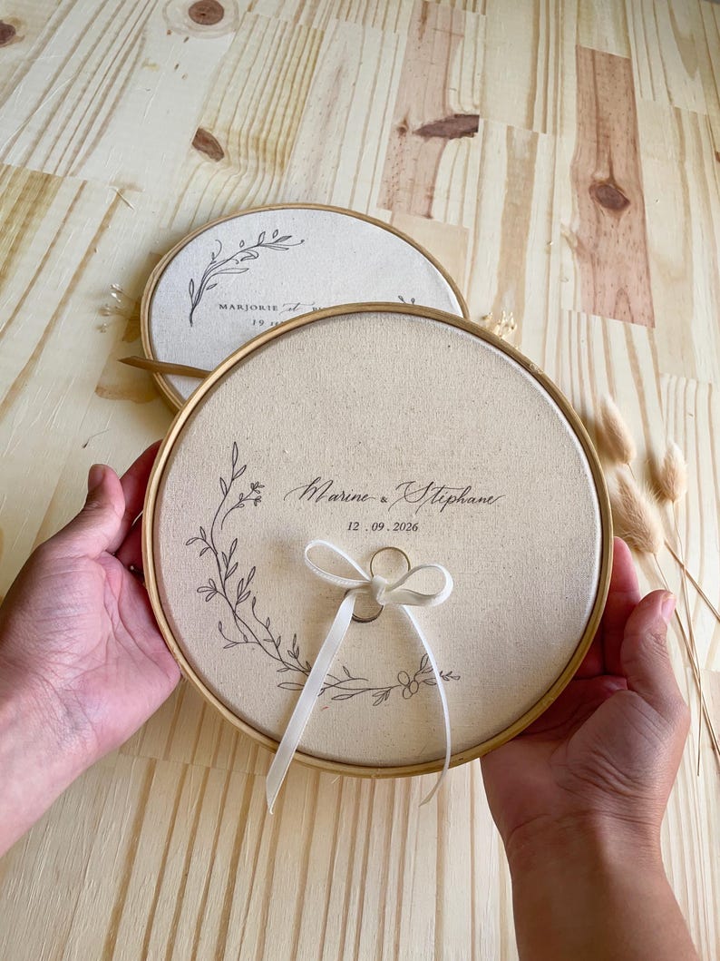 May include: Two round, beige ring holders with wooden frames. One features the names "Marine & Stephane" and the date "12.09.2026". The other has the name "Marjorie" and a date. Both have floral designs and a ribbon.