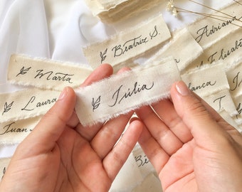 May include: A collection of small, rectangular, cream-colored fabric tags with black calligraphy. The tags have frayed edges and feature names written in cursive, including Alejandra, Beatriz S., Júlia, Marta, Laura, Juan, Adriana, Lisabe, Lanca, and Alberto.