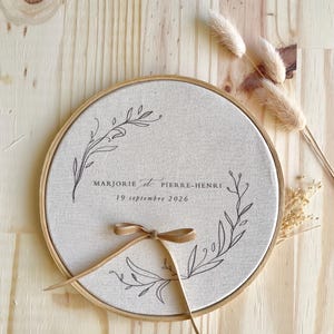 May include: A round, beige fabric embroidery hoop with a wooden frame. The fabric is printed with the names "Marjorie et Pierre-Henri" and the date "19 septembre 2026" in black script. A brown ribbon is tied in a bow at the bottom.