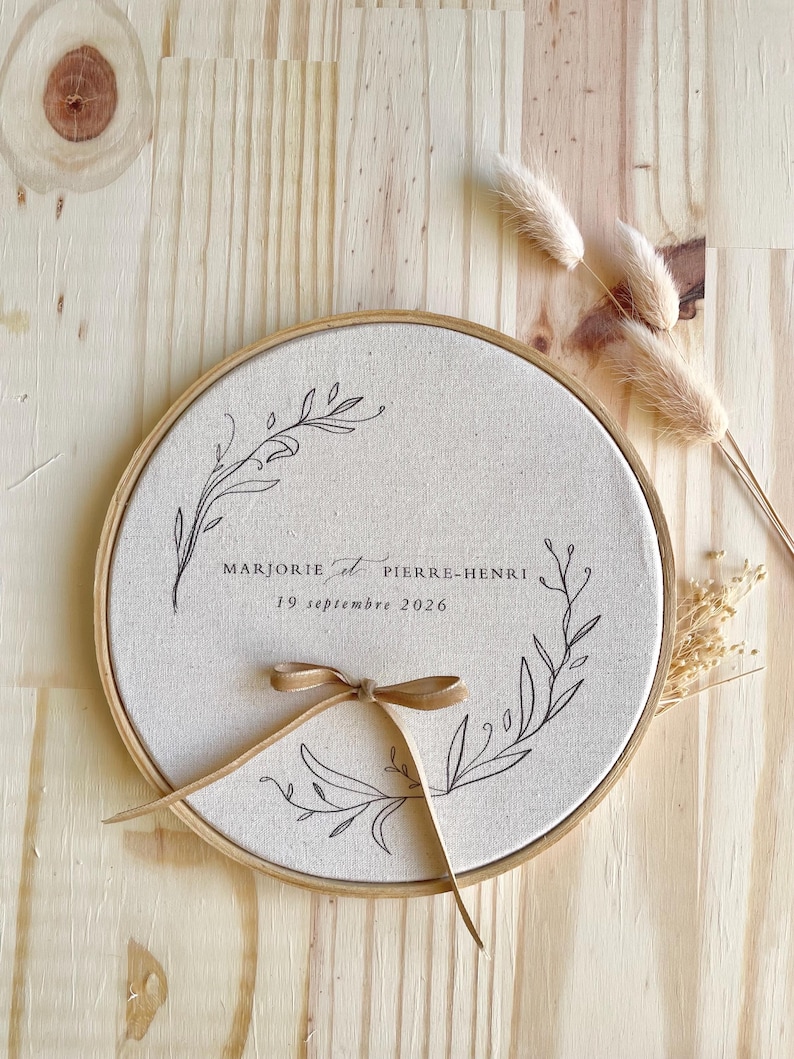 May include: A round, beige fabric embroidery hoop with a wooden frame. The fabric is printed with the names "Marjorie et Pierre-Henri" and the date "19 septembre 2026" in black script. A brown ribbon is tied in a bow at the bottom.