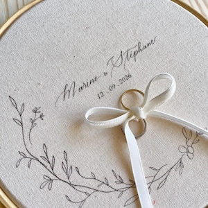 May include: A round, cream-colored ring bearer pillow with a wooden embroidery hoop frame. The fabric is printed with the names "Marine & Stephane" and the date "12.09.2026". A white ribbon holds two rings, and a floral design decorates the fabric.