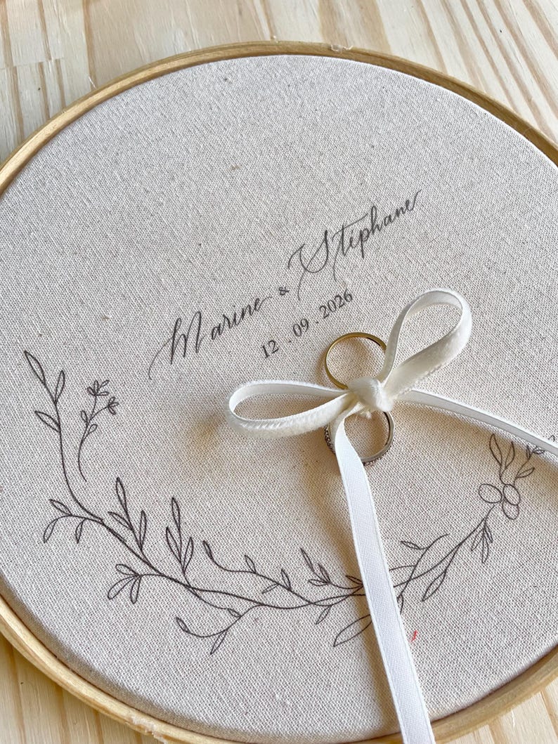 May include: A round, cream-colored ring bearer pillow with a wooden embroidery hoop frame. The fabric is printed with the names "Marine & Stephane" and the date "12.09.2026". A white ribbon holds two rings, and a floral design decorates the fabric.