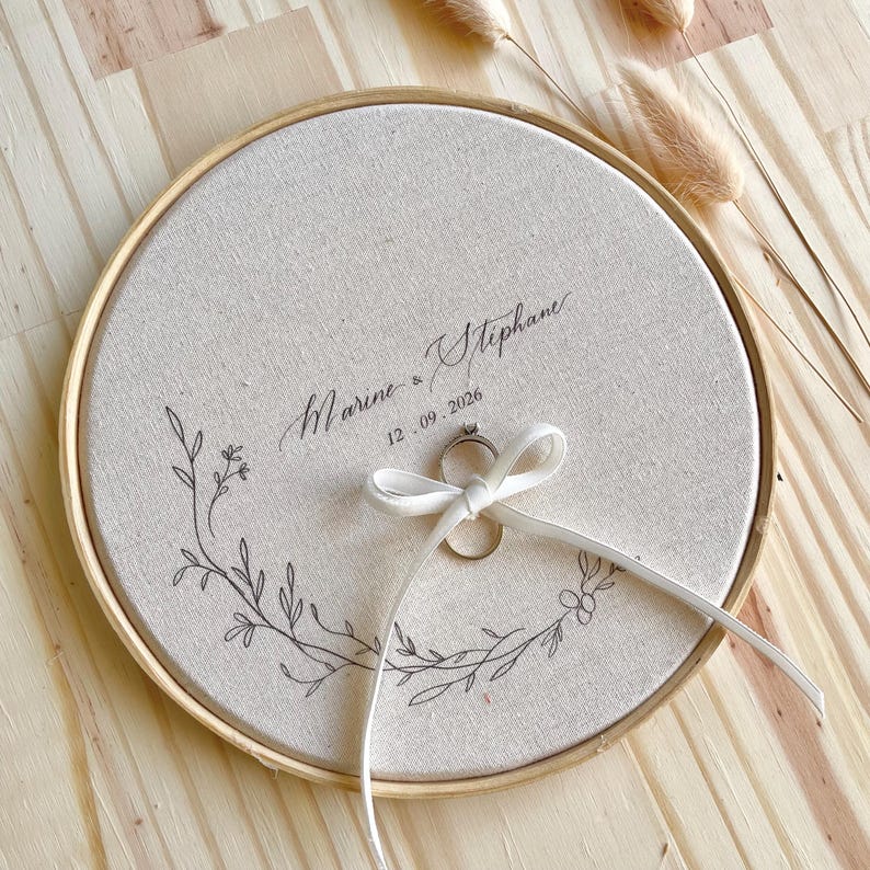 May include: A round, beige fabric ring holder with a wooden frame. The fabric is embroidered with a floral design and the names "Marine & Stephane" and the date "12.09.2026". A white ribbon holds a ring.