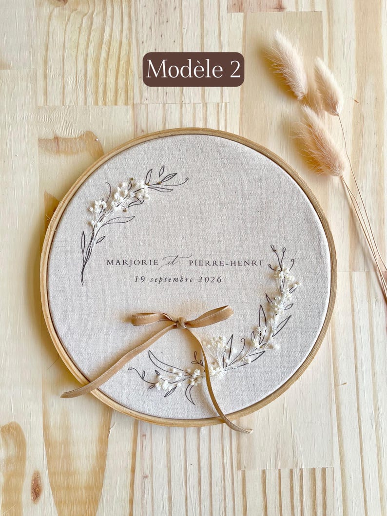 May include: A round, beige fabric embroidery hoop with a floral design and the names "MARJORIE et PIERRE-HENRI" and the date "19 septembre 2026" in black script. A tan ribbon is tied at the bottom. The text "Mod&egrave;le 2" is in a brown rectangle.