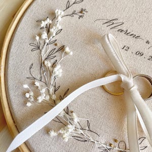 May include: A beige fabric ring bearer hoop with floral embroidery and the name "Marine" and a date. The hoop holds two gold wedding rings and a white ribbon. The embroidery features delicate white flowers and black line art.