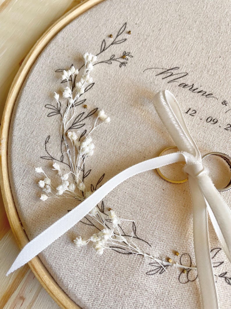 May include: A beige fabric ring bearer hoop with floral embroidery and the name "Marine" and a date. The hoop holds two gold wedding rings and a white ribbon. The embroidery features delicate white flowers and black line art.