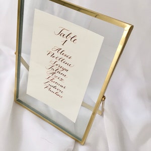 May include: A gold-framed glass table number holder with a white card that reads "Table 1" and lists the names Alexis, No&euml;lline, Soreya, Jatan, Alix, Fritz, Thomas, Ludovic, and Pauline in cursive.