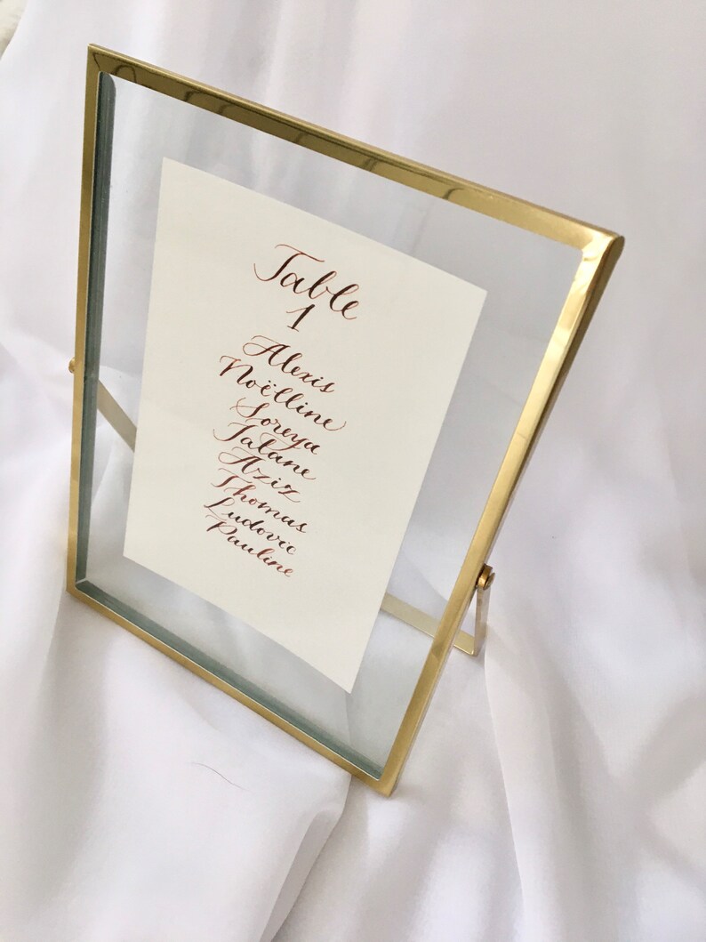 May include: A gold-framed glass table number holder with a white card that reads "Table 1" and lists the names Alexis, No&euml;lline, Soreya, Jatan, Alix, Fritz, Thomas, Ludovic, and Pauline in cursive.