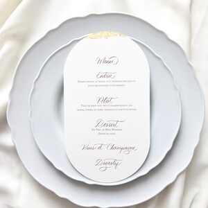 May include: A white oval menu card with gold accents and brown text. The menu includes the following sections: Entrie, Plat, Dessert, Vins et Champagne, Digestif. The text reads: "Menu Entrie Tomate rougie au soleil avec asperges grillees en sauce hollandaise et balsamique Plat Filet de boeuf avec sauce champignons et vin rouge, pomme de terre fondante et carottes botties Dessert Un trio de mini desserts Fruits en folie Vins et Champagne Digestif"