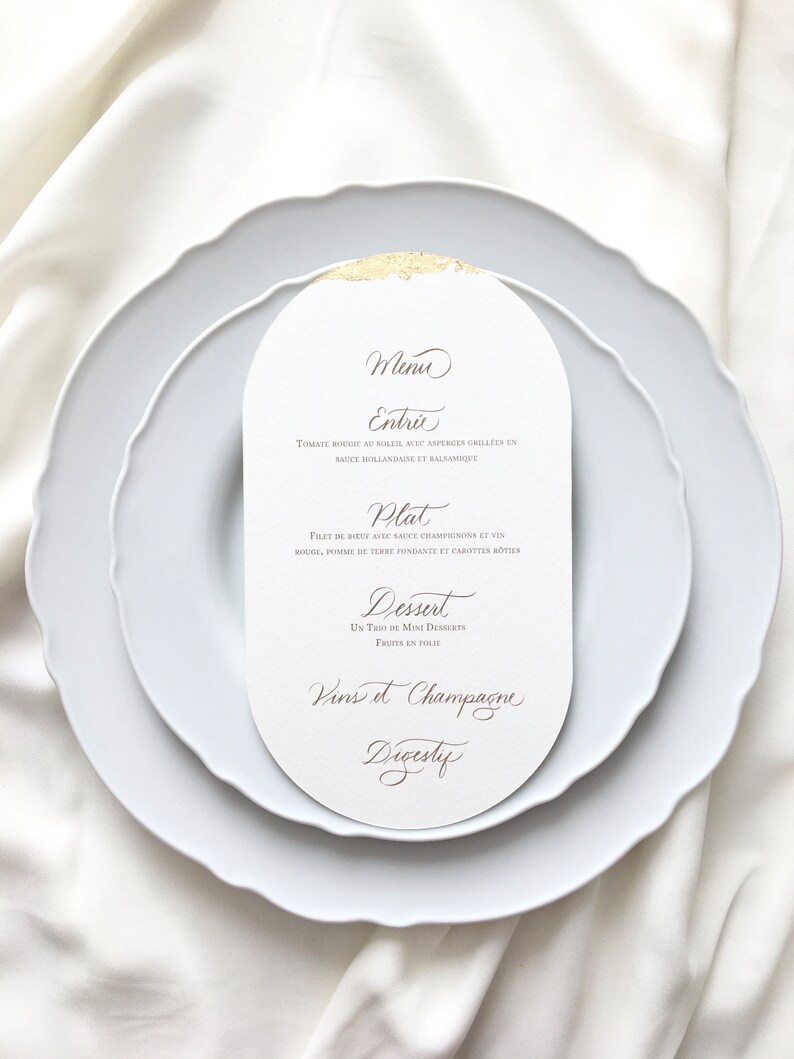May include: A white oval menu card with gold accents and brown text. The menu includes the following sections: Entrie, Plat, Dessert, Vins et Champagne, Digestif. The text reads: "Menu Entrie Tomate rougie au soleil avec asperges grillees en sauce hollandaise et balsamique Plat Filet de boeuf avec sauce champignons et vin rouge, pomme de terre fondante et carottes botties Dessert Un trio de mini desserts Fruits en folie Vins et Champagne Digestif"