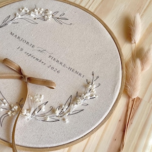Op de afbeelding: Een geborduurd huwelijkssouvenir met de namen "Marjorie et Pierre-Henri" en de datum "19 september 2026". Het ontwerp omvat bloemaccenten, een bruin lint en een houten hoepel. Gedroogde bloemen zijn rechts geplaatst.