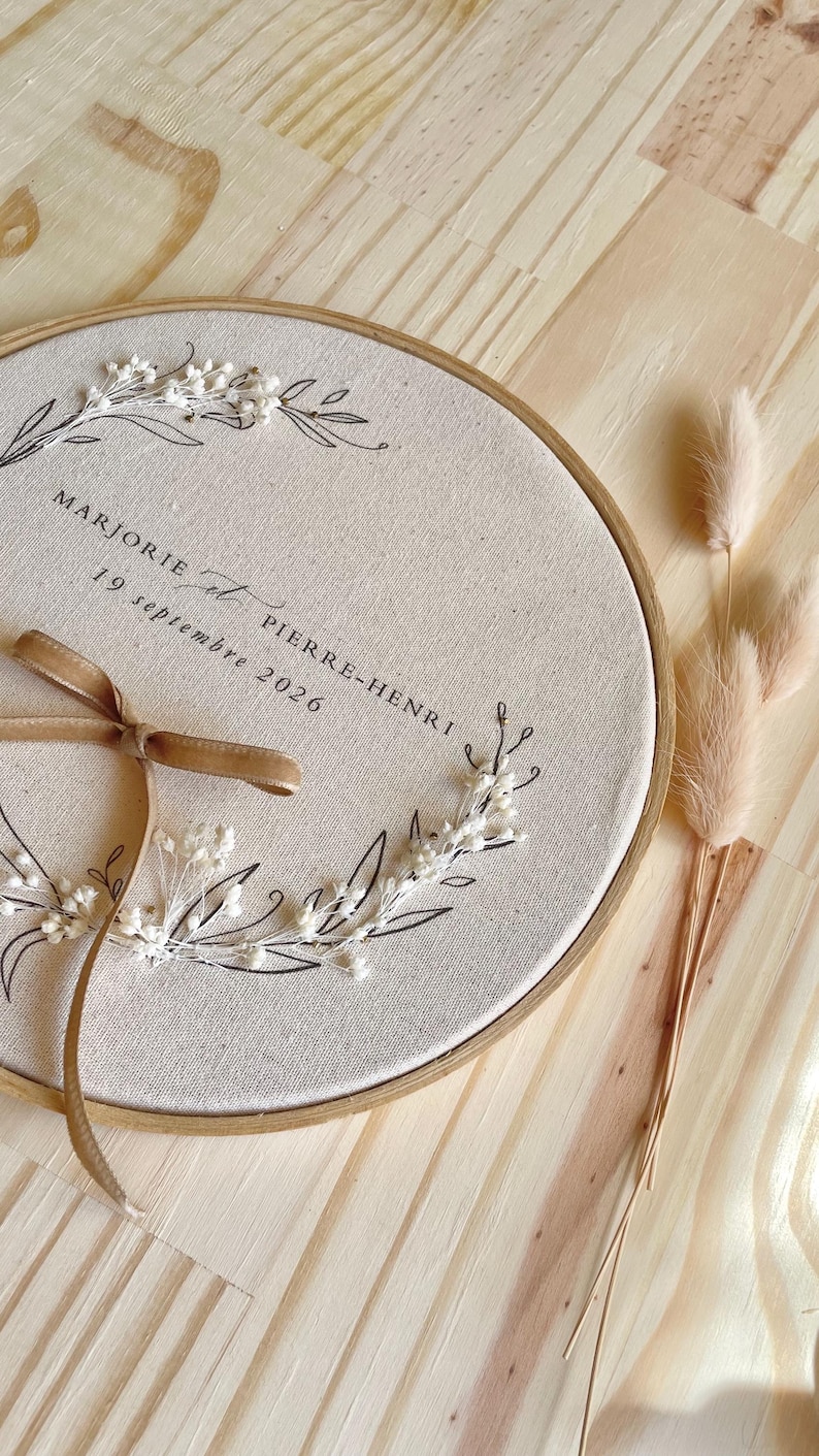 May include: A round, embroidered wedding keepsake featuring the names "Marjorie et Pierre-Henri" and the date "19 septembre 2026". The design includes floral accents, a brown ribbon, and a wooden hoop. Dried flowers are placed to the right.