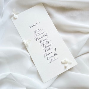 May include: A white card with black calligraphy listing the names "Elise, Florian, Benedicte, Kamul, Nelly, Olivier, Cedric, Jerome, Camille, Alexis" and the text "TABLE 1".