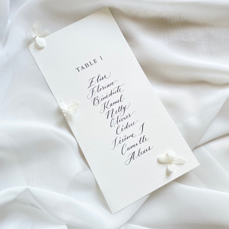 May include: A white card with black calligraphy listing the names "Elise, Florian, Benedicte, Kamul, Nelly, Olivier, Cedric, Jerome, Camille, Alexis" and the text "TABLE 1".