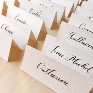 May include: A set of white place cards with black calligraphy names, including Chris, Louise, Dieter, Jean-Michel, Catherine, Guillaume, and Corinne. The cards are folded in half and arranged in a row on a wooden surface.