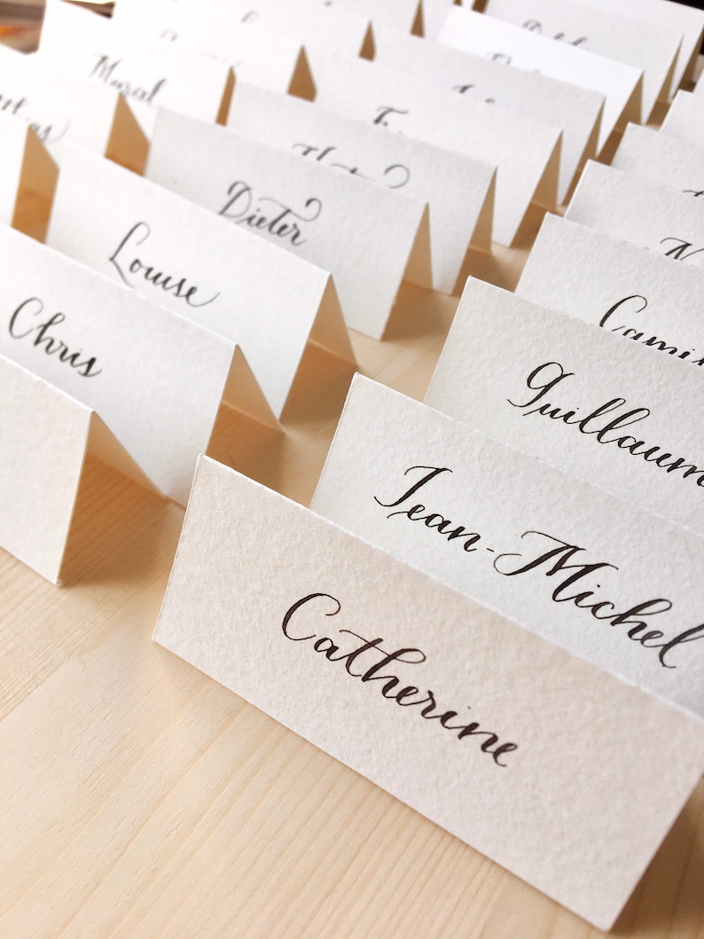 May include: A set of white place cards with black calligraphy names, including Chris, Louise, Dieter, Jean-Michel, Catherine, Guillaume, and Corinne. The cards are folded in half and arranged in a row on a wooden surface.