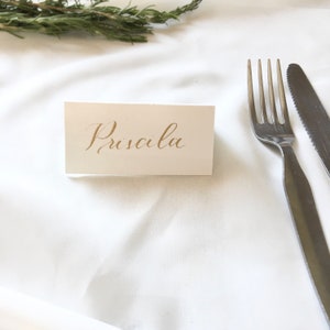 May include: A white place card with the name "Priscilla" written in gold calligraphy on a white tablecloth with a fork and knife.