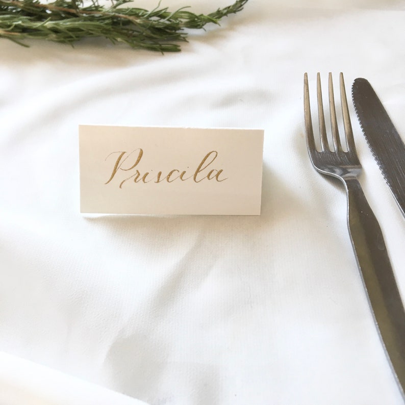 May include: A white place card with the name "Priscilla" written in gold calligraphy on a white tablecloth with a fork and knife.