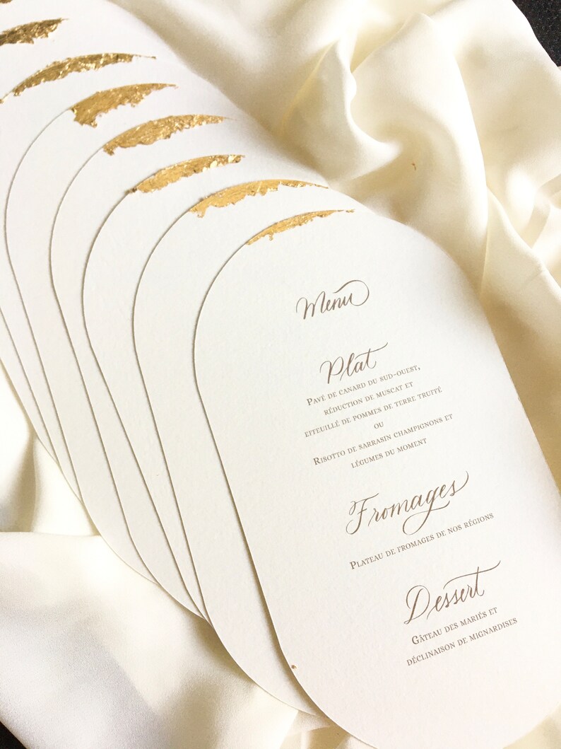 May include: A stack of white, oval-shaped cards with gold accents. The cards feature a French menu with the headings "Menu", "Plat", "Fromages", and "Dessert".