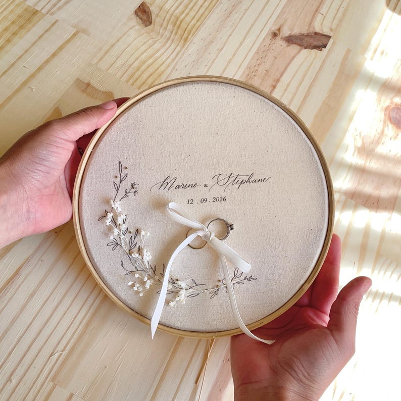 May include: A round, cream-colored ring bearer pillow with a wooden frame. The pillow features the names "Marine & Stephane" and the date "12.09.2026" in elegant script, with a floral design. Two rings are tied with a white ribbon.