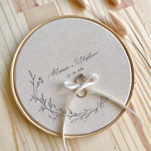 May include: A round, beige fabric ring holder with a wooden frame. The fabric is embroidered with a floral design and the names "Marine & Stephane" and the date "12.09.2026". A white ribbon holds a ring.