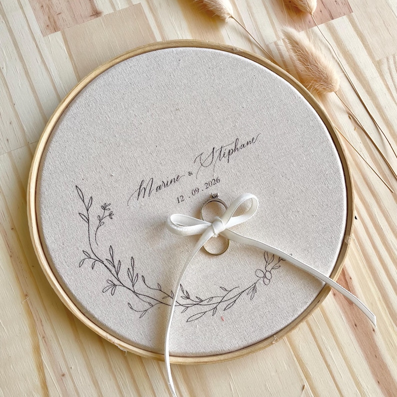 May include: A round, beige fabric ring holder with a wooden frame. The fabric is embroidered with a floral design and the names "Marine & Stephane" and the date "12.09.2026". A white ribbon holds a ring.