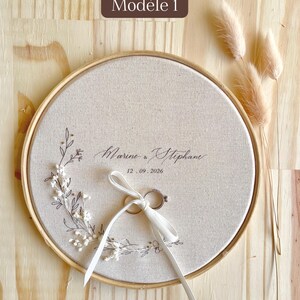 May include: A round, beige ring bearer pillow with a wooden frame. The pillow features the names "Marine & Stephane" and the date "12.09.2026" in elegant script, with a white ribbon holding two rings. Decorative floral elements and the text "Mod&egrave;le 1" are also visible.