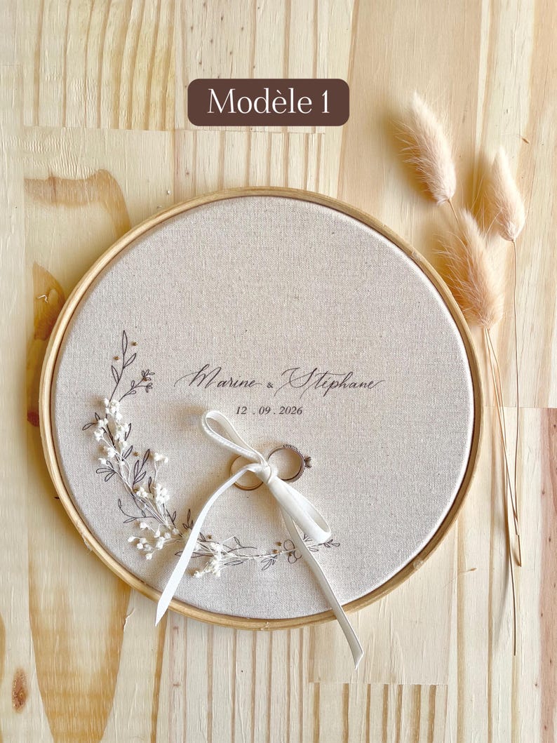 May include: A round, beige ring bearer pillow with a wooden frame. The pillow features the names "Marine & Stephane" and the date "12.09.2026" in elegant script, with a white ribbon holding two rings. Decorative floral elements and the text "Mod&egrave;le 1" are also visible.
