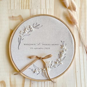 May include: A round, beige fabric embroidery hoop with the names "MARJORIE et PIERRE-HENRI" and the date "19 septembre 2026" in black script. The design includes floral elements and a tan ribbon bow. The background is a light wood surface.
