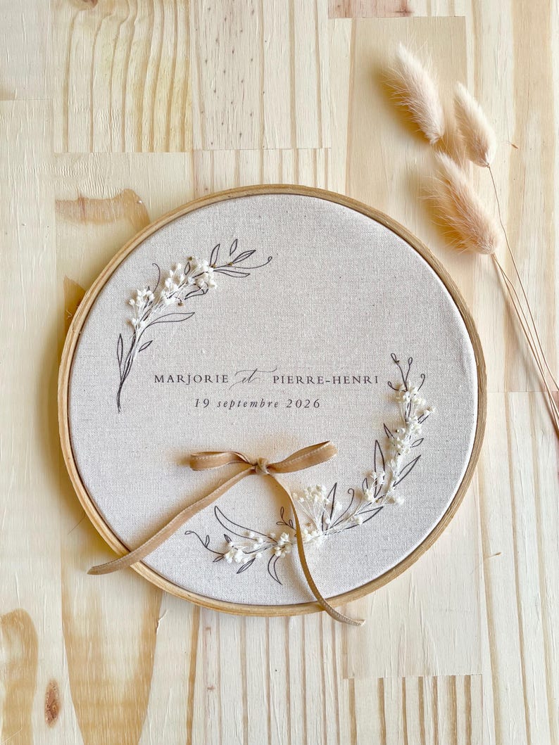 May include: A round, beige fabric embroidery hoop with the names "MARJORIE et PIERRE-HENRI" and the date "19 septembre 2026" in black script. The design includes floral elements and a tan ribbon bow. The background is a light wood surface.