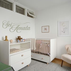 May include: A nursery room with a white crib, changing table, and a cozy armchair. The crib has light pink bedding. A framed print of a bunny and moon hangs on the wall. The wall has the text "Sei Amata" in cursive.