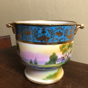 May include: A white porcelain vase with gold accents and a blue and gold band around the top. The vase has a landscape scene painted on the side with a house, trees, and a lake. The vase has two gold handles on the sides.