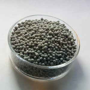 May include: A clear glass petri dish filled with numerous small, round, gray spheres. The spheres are densely packed, filling the dish. The dish is set against a white background, providing contrast to the gray spheres.