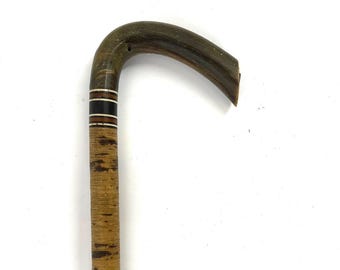 May include: A vintage wooden cane with a curved handle. The handle is dark brown, and the shaft is a lighter brown with dark brown accents. A decorative band of white, black, and brown stripes is near the handle.
