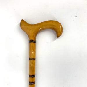 Puede incluir: Un bastón de madera con un mango curvo. El bastón es de madera marrón claro con bandas marrones oscuras cerca de la parte superior. El mango es liso y pulido, proporcionando un agarre cómodo. El bastón está diseñado para soporte y equilibrio.