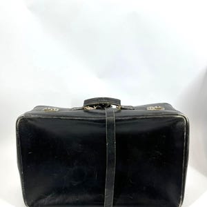 Op de afbeelding: Een zwarte vintage koffer met een leren riem en metalen sluitingen. De rechthoekige koffer heeft een handvat aan de bovenkant en metalen hardware. De buitenkant vertoont tekenen van slijtage.