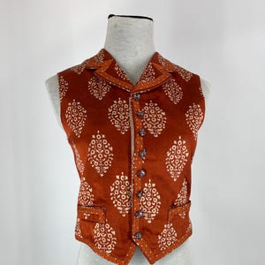 May include: A sleeveless waistcoat in a burnt orange hue featuring a repeating white floral block print pattern. The garment includes a notched lapel collar, a front button closure with six dark circular buttons, and two small welt pockets at the waist.