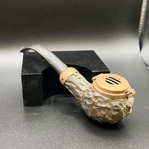 Peut inclure: Une pipe à tabac en bois sculpté avec un tuyau sombre et incurvé et un couvercle métallique articulé. Le foyer de la pipe a un aspect texturé en forme d'écorce, et le couvercle comporte trois fentes d'aération. La pipe repose sur une surface noire.