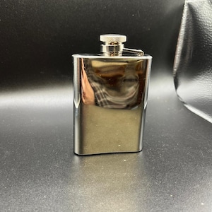 May include: A rectangular stainless steel hip flask with a polished, mirror-like finish. The flask features a rounded bottom edge and a secure, screw-top cap with a textured, knurled grip. The metal surface reflects the surrounding environment.