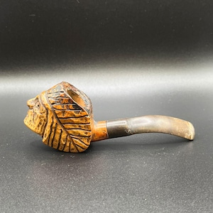 May include: A carved wooden tobacco pipe featuring a leaf and face design on the bowl. The pipe has a dark brown stem and a metal band. The wood is a warm, natural colour. The pipe is approximately 15 cm long.