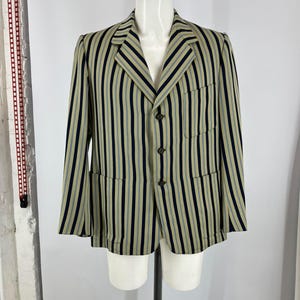 May include: A blazer featuring a vertical striped pattern in tan, navy blue, and light blue. The garment has a notched lapel, three dark buttons down the front, one chest pocket, and two lower pockets. The jacket is displayed on a mannequin torso.