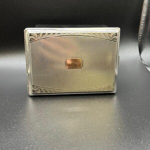 May include: A rectangular silver-toned metal cigarette case featuring a fine grid-patterned texture across the front surface. The case includes a smooth, polished rectangular center plaque and decorative etched leaf-like flourishes at the corners.
