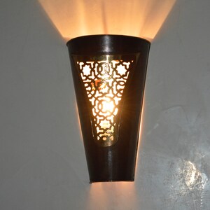 Op de afbeelding: Een zwarte metalen wandlamp met een uitgesneden messing ontwerp en een warm licht dat erdoorheen schijnt.