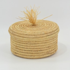 May include: A round, natural woven basket with a lid and a small, natural fibre tassel on top.