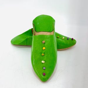 Pode incluir: Chinelos de couro verde brilhante com bico fino. Os chinelos são decorados com pequenos e coloridos pompons. Estes chinelos feitos à mão são de cor verde brilhante e provavelmente para uso interno.