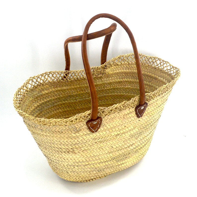 K&ouml;nnte beinhalten: Eine gro&szlig;e, ovale Strohm&auml;rkttasche mit braunen Ledergriffen. Die Tasche hat ein gewebtes Design und einen geh&auml;kelten Besatz am oberen Rand.