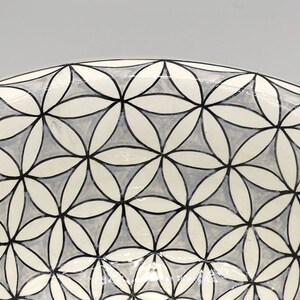 May include: A ceramic bowl with a geometric floral pattern. The design features interlocking flower shapes in white, light gray, and black. The rim of the bowl is a solid gray color. The bowl is likely used for serving or display.