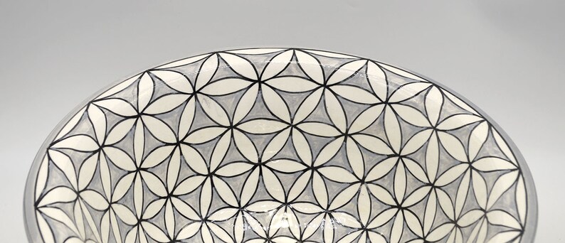 May include: A ceramic bowl with a geometric floral pattern. The design features interlocking flower shapes in white, light gray, and black. The rim of the bowl is a solid gray color. The bowl is likely used for serving or display.