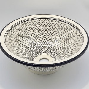 May include: Cream ceramic basin with a black rim and a detailed black fish scale pattern. The sink has a circular shape with a drain hole at the bottom. The design is symmetrical and decorative.